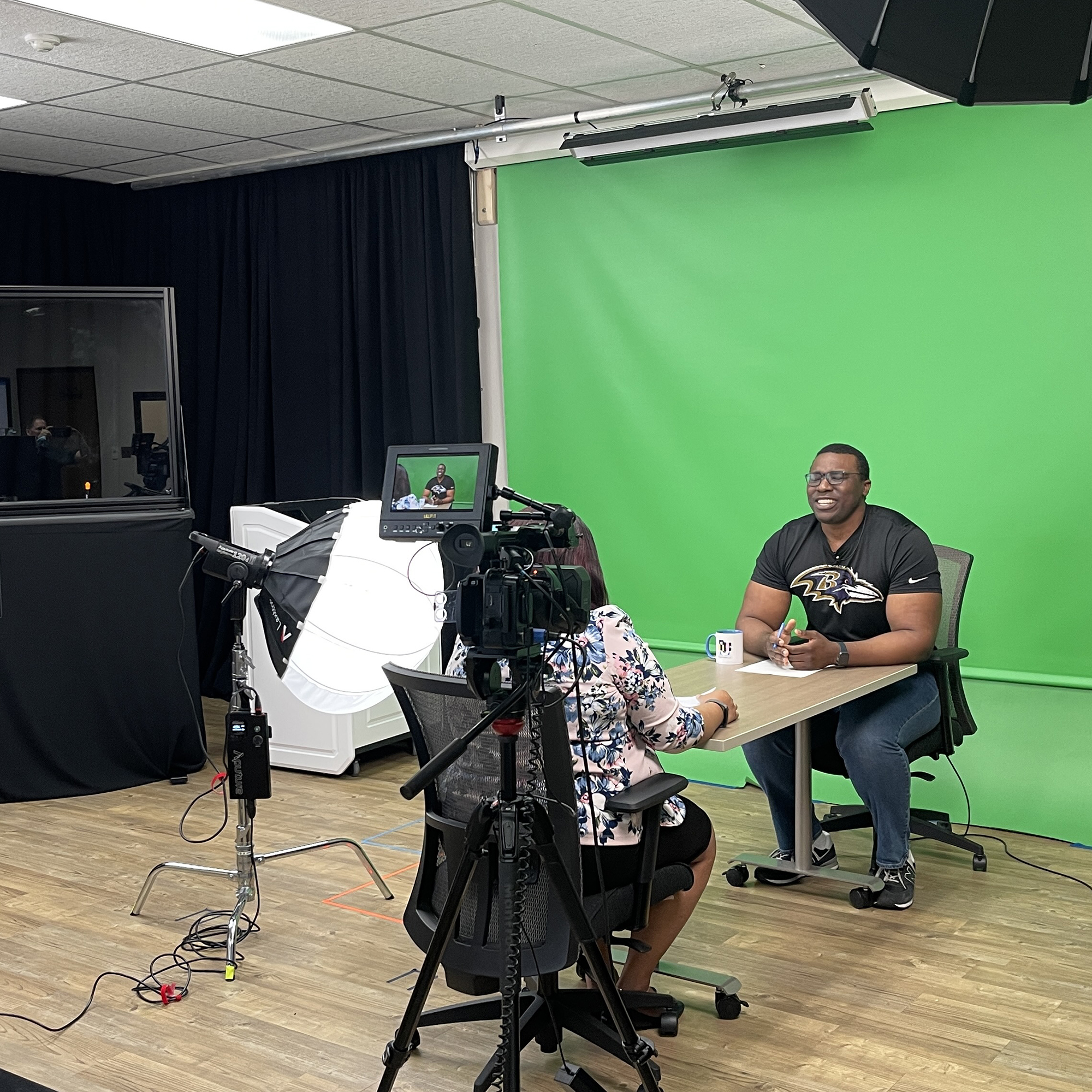 Two people sit at a desk in the Innovation Lab with a green screen behind one of the people