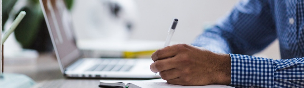 Person writing in a notebook at a desk with a laptop in the background