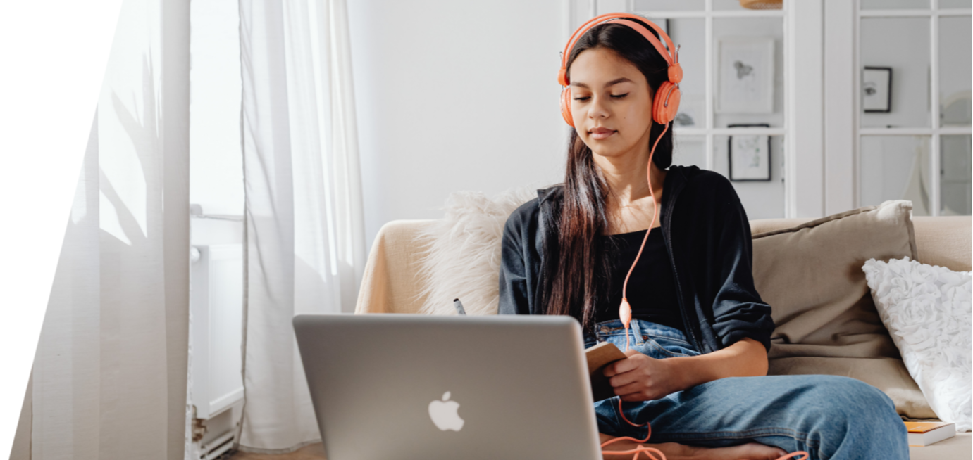 Photo of a student with long dark hair seated in front of a laptop, wearing headphones and writing in a notebook