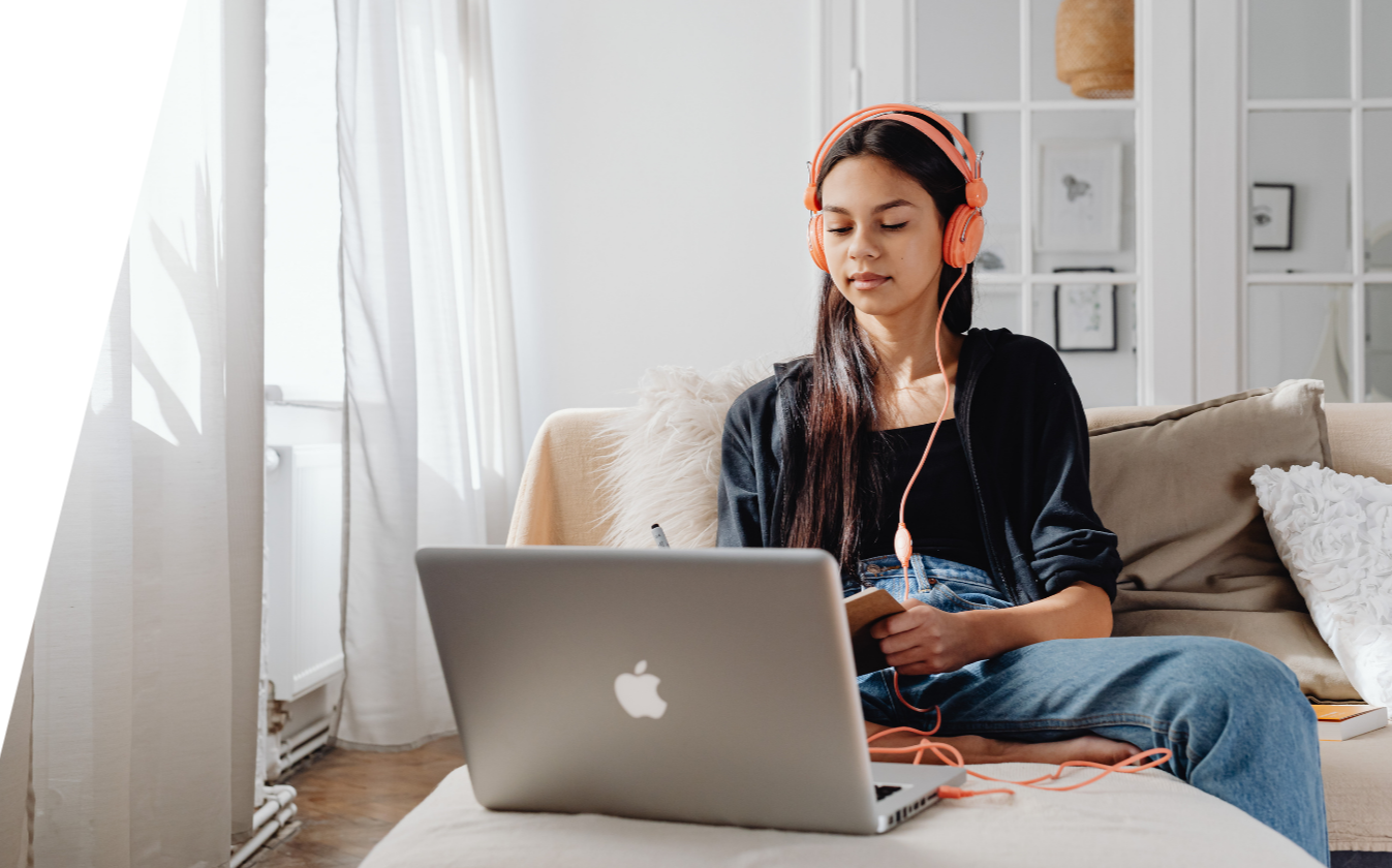 Photo of student with dark hair seated in front of a laptop, wearing headphones and writing in a notebook