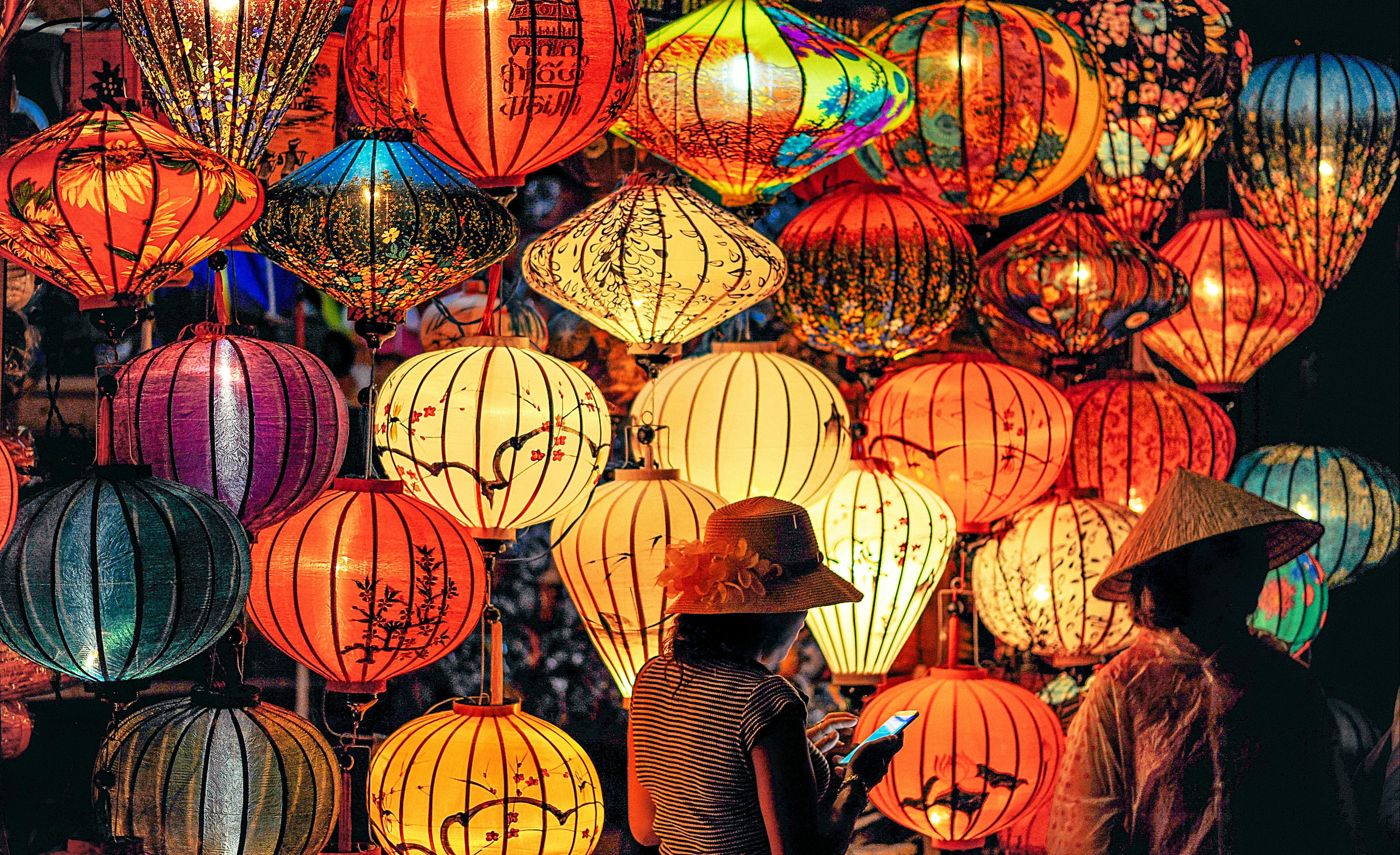 A photo of many colourful paper lanterns lit from inside, with two people wearing hats in the foreground