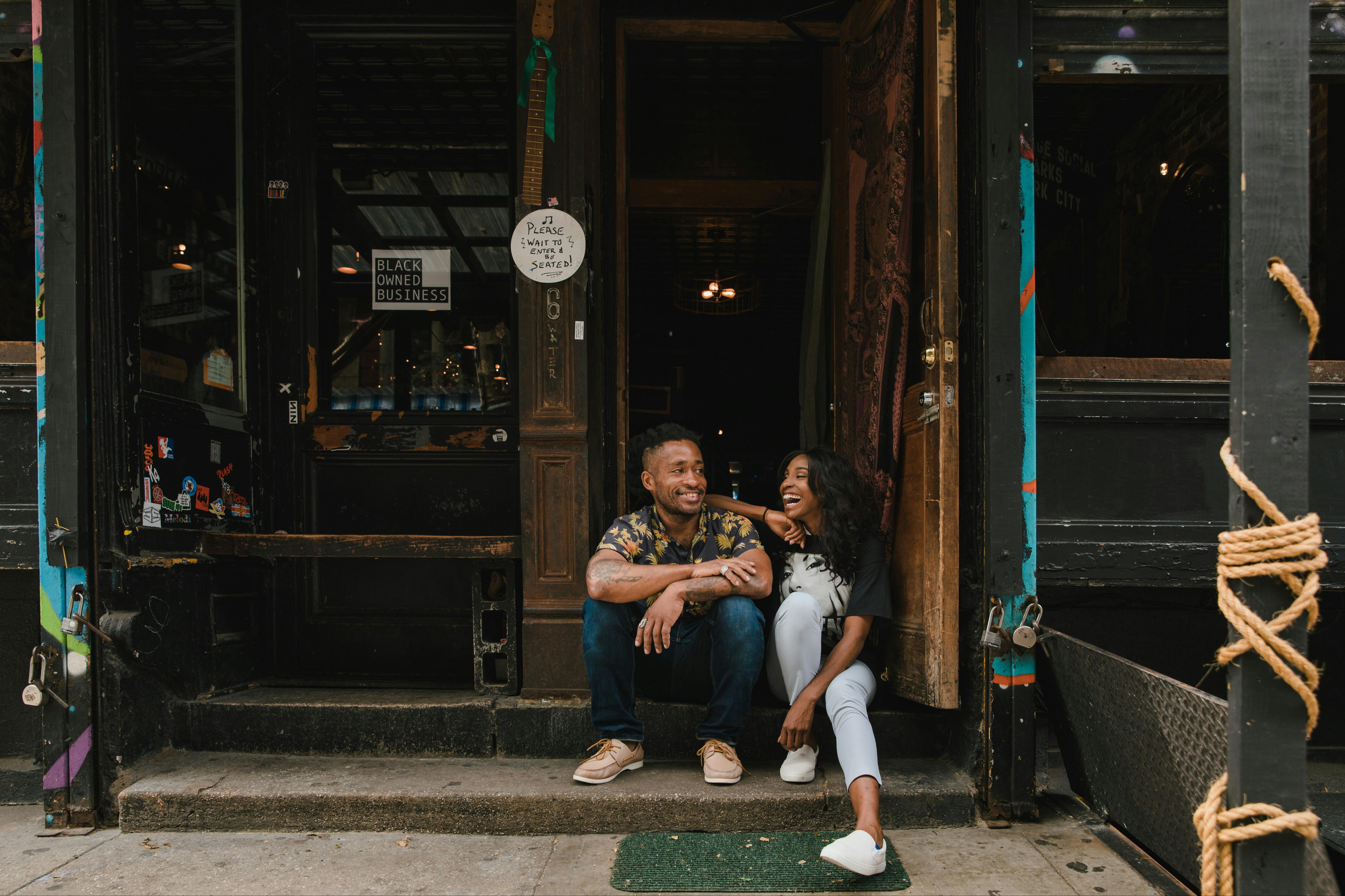 Photo of two Black people sitting on the stoop in front of a building, with the sign "Black Owned Business" in the window