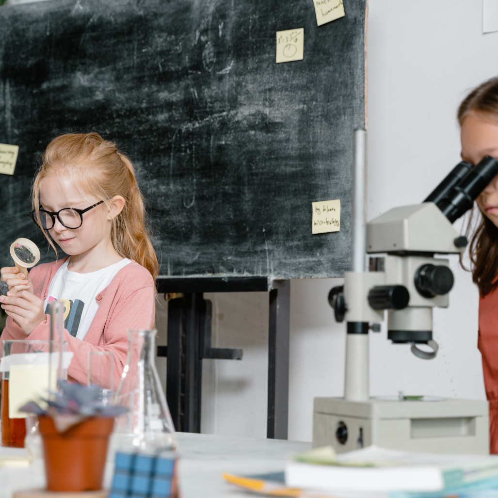 2 girls in a classroom , one is using a microscope the other a magnifying glass